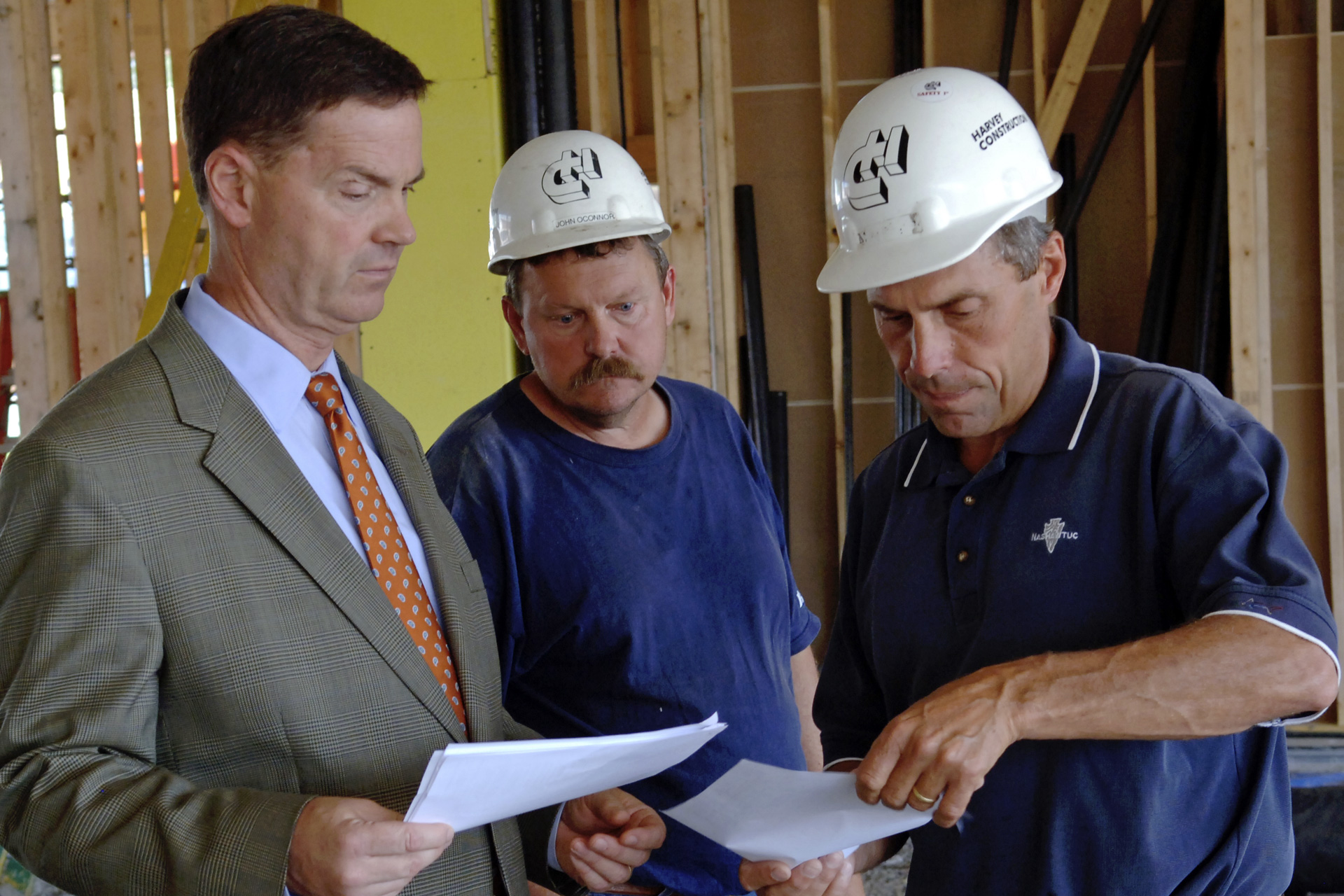 Man in suit and tie talks with a couple builders in safety helmets