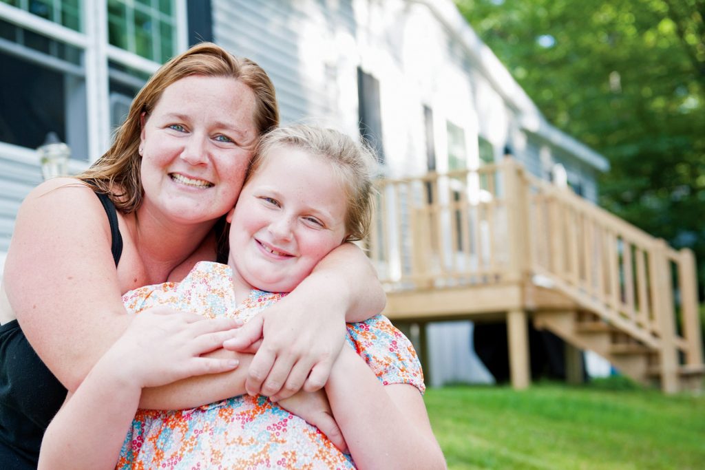 Woman hugging her daughter in their front yard, with a porch behind them.