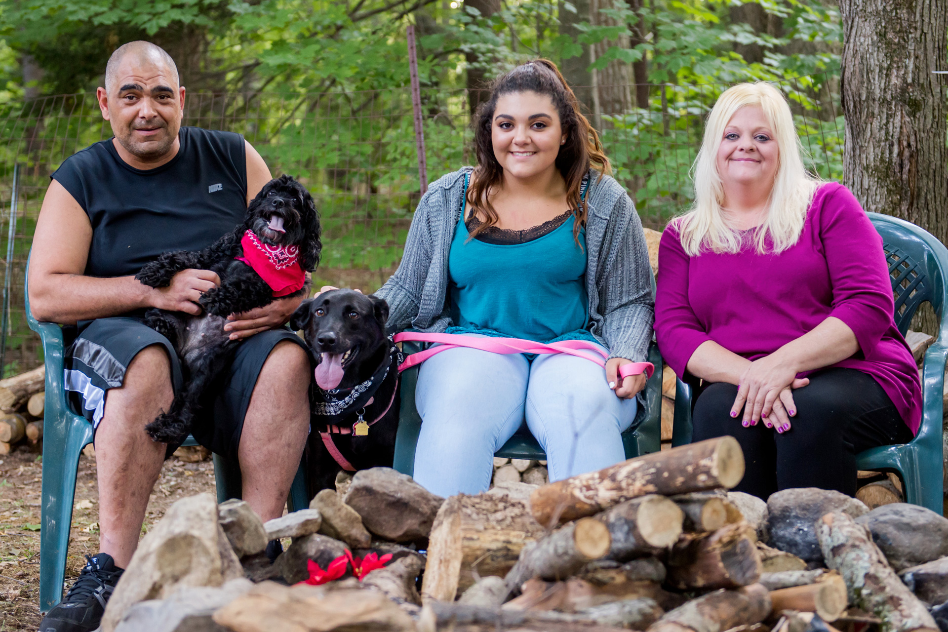 A dad, mom, daughter, and two dogs sit outdoors by the wood pile