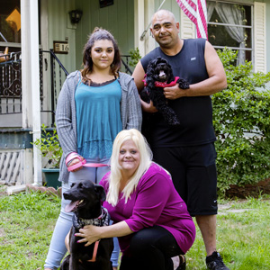 A dad, mom, daughter, and two dogs sit stand outside their manufactured home
